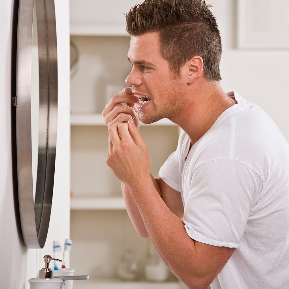 Bakteriell bedingtes Zahnfleischbluten kann durch konsequente Pflege zu Hause vorgebeugt werden. Ein Mann steht vor dem Spiegel und reinigt die Zahnzwischenräume mit Zahnseide.